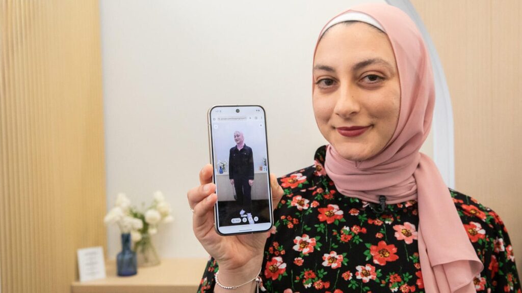 A woman in a pink headscarf and floral top smiles at the camera and holds a phone with an image of her wearing a denim jumpsuit